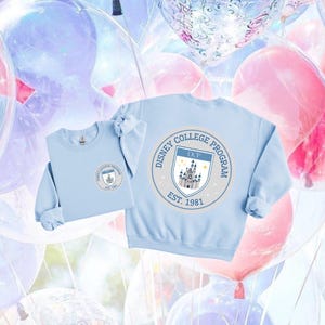 May include: A light blue crewneck sweatshirt featuring a circular graphic on the front left chest and a larger version on the back. The design includes the text Disney College Program, Est. 1981, and DCP above an illustration of a castle with stars.
