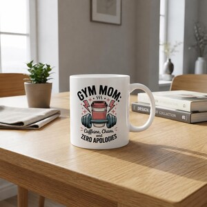 May include: White ceramic mug with the text "GYM MOM: Caffeine, Chaos, and Zero Apologies." The mug features a graphic of a coffee cup, dumbbells, and lightning bolts. The mug is on a wooden table.