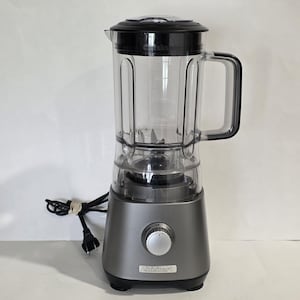 May include: A Cuisinart blender with a clear plastic pitcher and a black lid and handle. The base is gray with a control dial and the Cuisinart logo. The power cord is visible.