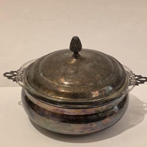 May include: A silver-plated serving dish with a glass insert and a decorative lid. The lid is topped with a pinecone-shaped finial. The dish has ornate handles and a reflective, iridescent finish.