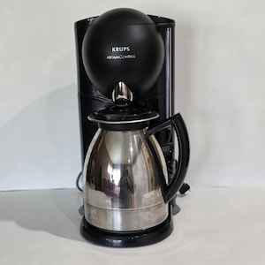 May include: A black KRUPS Aroma Control coffee maker with a stainless steel thermal carafe. The coffee maker has a round top and a black handle on the carafe. The text "KRUPS AromaControl" is printed on the front.