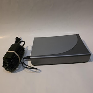 Puede incluir: Un dispositivo electrónico gris con una parte superior curva y un adaptador de corriente negro. El dispositivo tiene forma rectangular con una esquina redondeada. El adaptador de corriente es negro con una banda blanca.