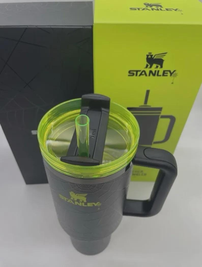 Puede incluir: Un vaso Stanley negro con un patr&oacute;n de telara&ntilde;a, una tapa transl&uacute;cida de color verde lima, una pajita a juego de color verde lima y un asa negra. El vaso presenta el logotipo de Stanley en verde lima. Est&aacute; colocado frente a su caja verde lima y negra.