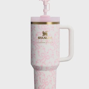May include: A white Stanley tumbler with a pink ribbon and floral pattern, featuring a gold logo and the text LOVESHACKFANCY. The tumbler has a side handle, a pink lid, and a straw topper shaped like a pink bow, all set against a plain background.