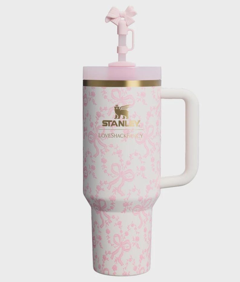May include: A white Stanley tumbler with a pink ribbon and floral pattern, featuring a gold logo and the text LOVESHACKFANCY. The tumbler has a side handle, a pink lid, and a straw topper shaped like a pink bow, all set against a plain background.