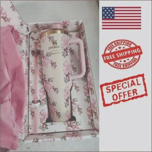 May include: A cream-colored Stanley tumbler with a floral pattern featuring pink and dark red flowers, a gold-tone rim, and a matching handle. The tumbler is presented in a box with a coordinating floral print, accompanied by a clear plastic straw.