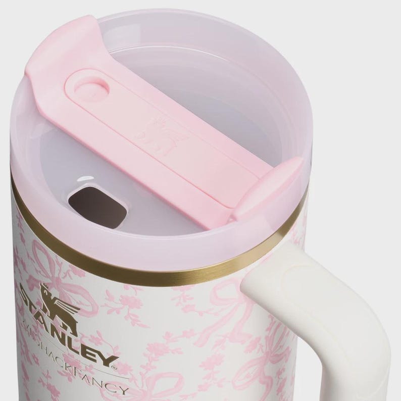 May include: A white insulated tumbler featuring a pink floral and ribbon pattern with a gold-tone rim. The lid is translucent with a light pink sliding closure. The Stanley logo and LoveShackFancy text are printed on the side in black ink.