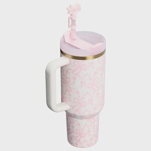 May include: A white insulated tumbler featuring a repeating light pink bow and floral pattern. The cup includes a sturdy white side handle, a gold-toned rim, a light pink lid, and a matching pink straw topped with a decorative bow accessory.