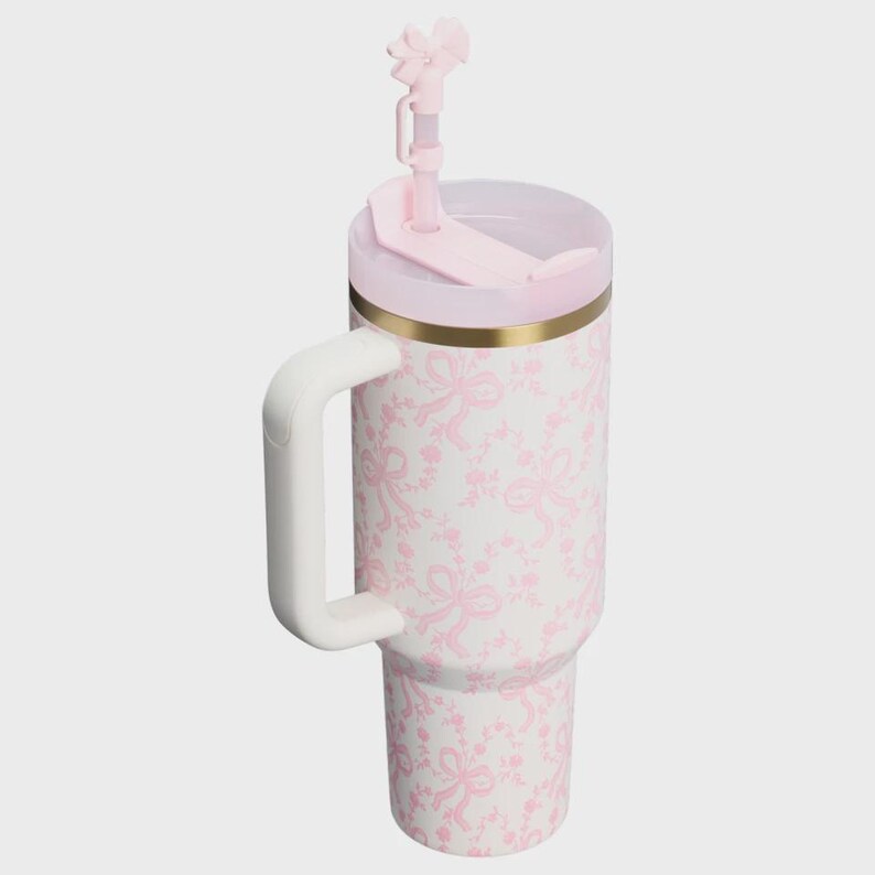 May include: A white insulated tumbler featuring a repeating light pink bow and floral pattern. The cup includes a sturdy white side handle, a gold-toned rim, a light pink lid, and a matching pink straw topped with a decorative bow accessory.