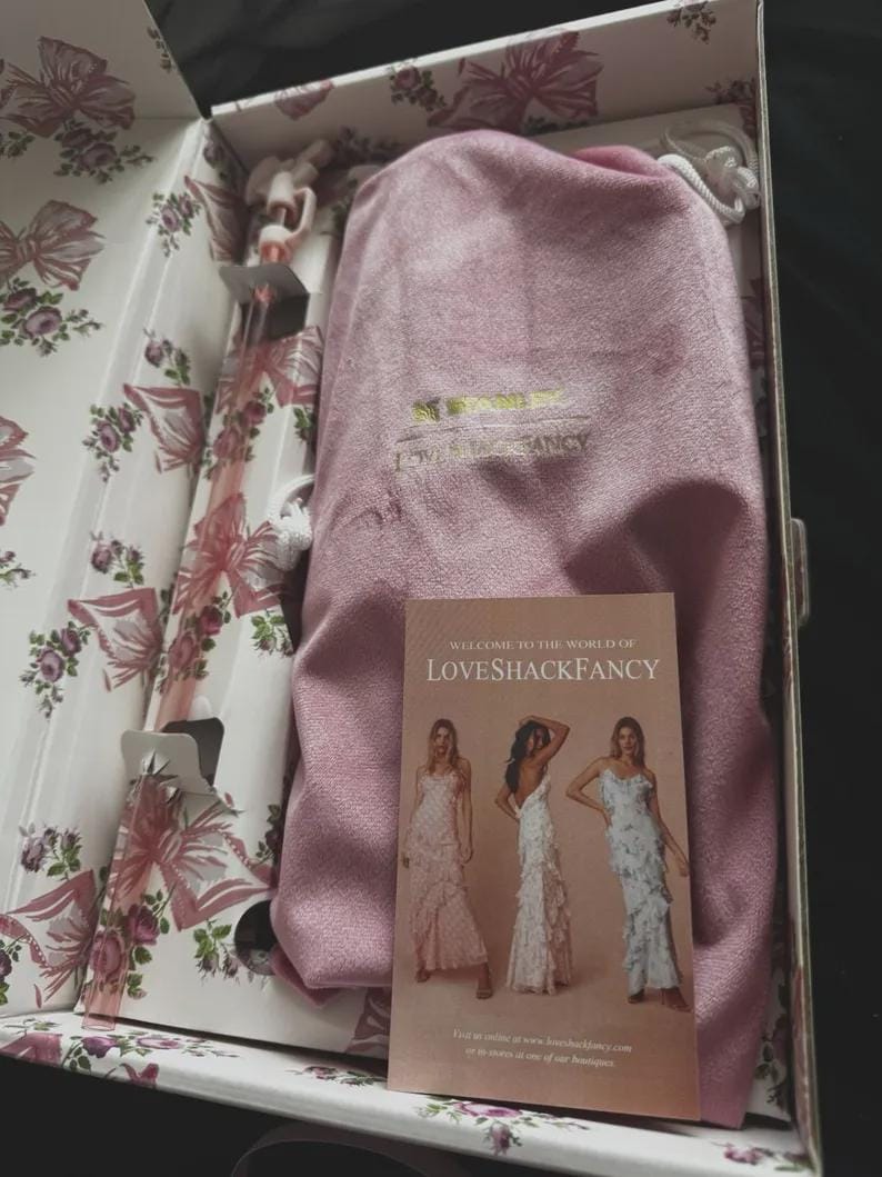 May include: A pink velvet drawstring pouch with gold lettering inside a floral-patterned box. The box contains a clear glass straw with a pink bow detail and a rectangular card featuring the text Welcome to the world of LoveShackFancy.