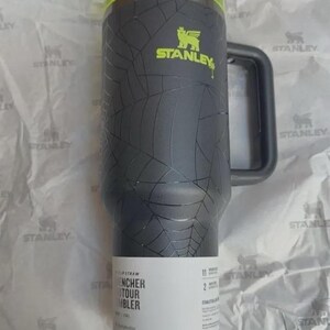 Puede incluir: Un vaso Stanley negro con un patr&oacute;n de telara&ntilde;a, una tapa de color amarillo ne&oacute;n y un asa gris oscuro. El vaso presenta el logotipo de Stanley en amarillo ne&oacute;n y una etiqueta blanca en la base con texto que incluye "FLIP STRAW" y "QUENCHER H2.0 FLOWSTATE TUMBLER".