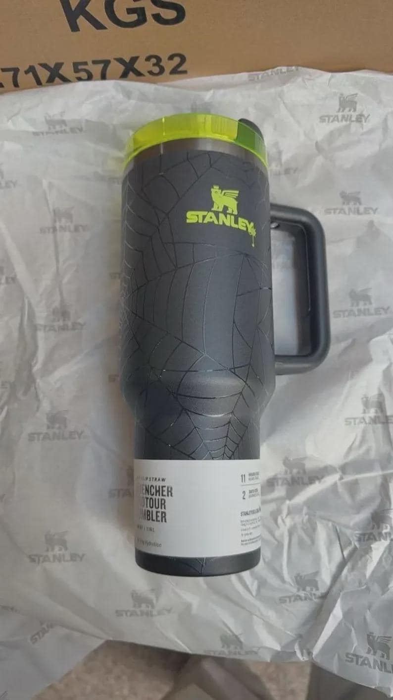 Puede incluir: Un vaso Stanley negro con un patr&oacute;n de telara&ntilde;a, una tapa de color amarillo ne&oacute;n y un asa gris oscuro. El vaso presenta el logotipo de Stanley en amarillo ne&oacute;n y una etiqueta blanca en la base con texto que incluye "FLIP STRAW" y "QUENCHER H2.0 FLOWSTATE TUMBLER".