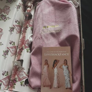 May include: A pink velvet drawstring pouch with gold lettering inside a floral-patterned box. The box contains a clear glass straw with a pink bow detail and a rectangular card featuring the text Welcome to the world of LoveShackFancy.