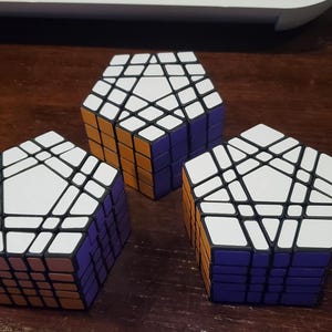 May include: Three dodecahedron-shaped twisty puzzles with white top faces, orange side panels, and purple lower sections. Each puzzle features a complex grid of smaller segments arranged in a pentagonal pattern, showcasing a multi-layered design.