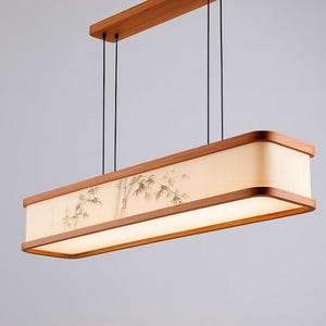 May include: Rectangular wooden pendant light fixture with a bamboo design on the shade. The light has a warm, soft glow and is suspended by black cords from a wooden ceiling mount. The fixture has a natural wood finish.