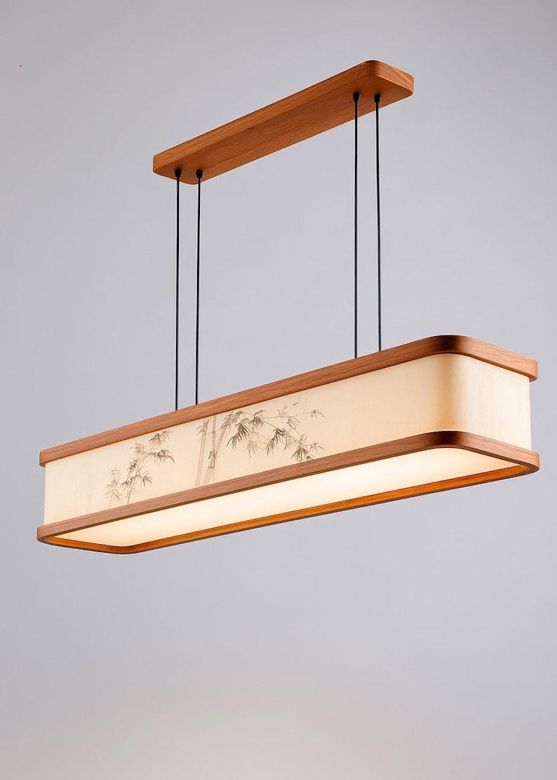 May include: Rectangular wooden pendant light fixture with a bamboo design on the shade. The light has a warm, soft glow and is suspended by black cords from a wooden ceiling mount. The fixture has a natural wood finish.