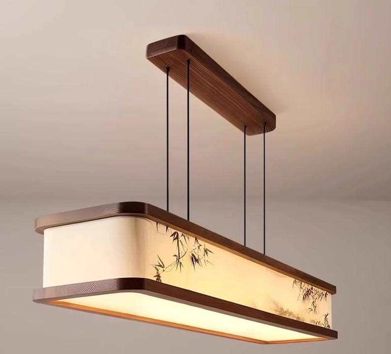 May include: Rectangular pendant light fixture with a wooden frame and a bamboo design on the shade. The light hangs from the ceiling by black cords. The fixture has a warm, inviting glow.
