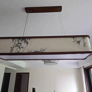 May include: Rectangular hanging light fixture with a dark wood frame and a cream-colored shade. The shade features a black bamboo design. The fixture is suspended from the ceiling by thin wires.