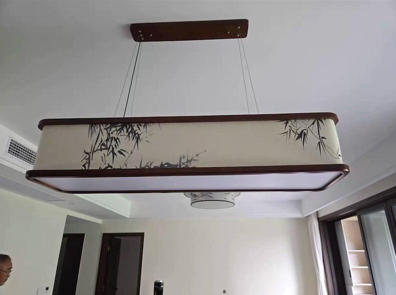 May include: Rectangular hanging light fixture with a dark wood frame and a cream-colored shade. The shade features a black bamboo design. The fixture is suspended from the ceiling by thin wires.