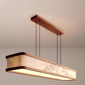 May include: A rectangular pendant light fixture with a wooden frame and a light-colored shade. The shade features a painted bamboo design. The fixture is suspended from the ceiling by thin black cords and a wooden beam.