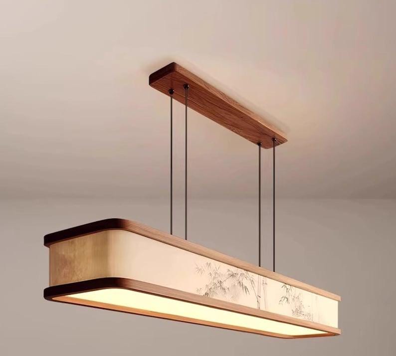 May include: A rectangular pendant light fixture with a wooden frame and a light-colored shade. The shade features a painted bamboo design. The fixture is suspended from the ceiling by thin black cords and a wooden beam.