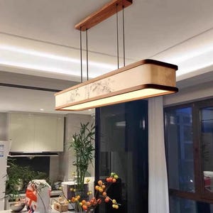 May include: Rectangular wooden pendant light fixture. The light has a light beige shade with a painted design and a warm glow. The fixture is suspended from the ceiling by black cords and a rectangular wooden mount.