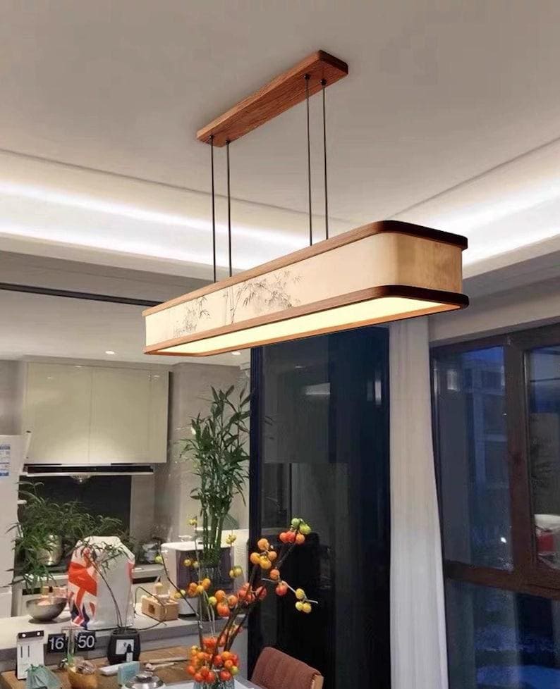 May include: Rectangular wooden pendant light fixture. The light has a light beige shade with a painted design and a warm glow. The fixture is suspended from the ceiling by black cords and a rectangular wooden mount.