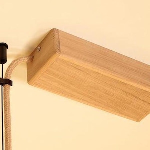 May include: A wooden rectangular light fixture with a natural finish is mounted on a wall. A beige fabric-covered cord and a black electrical component are attached. The fixture is angled, showcasing its wooden construction.