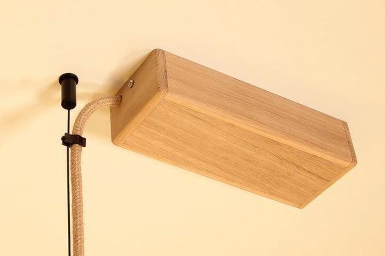 May include: A wooden rectangular light fixture with a natural finish is mounted on a wall. A beige fabric-covered cord and a black electrical component are attached. The fixture is angled, showcasing its wooden construction.