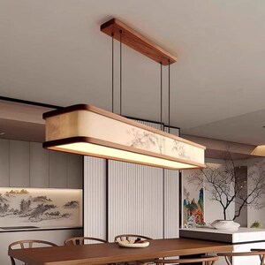 May include: A rectangular, wooden pendant light fixture with a warm, ambient glow. The fixture is suspended from the ceiling by thin black cords and features a light-colored shade with a subtle, Asian-inspired landscape design. The fixture is above a wooden dining table.
