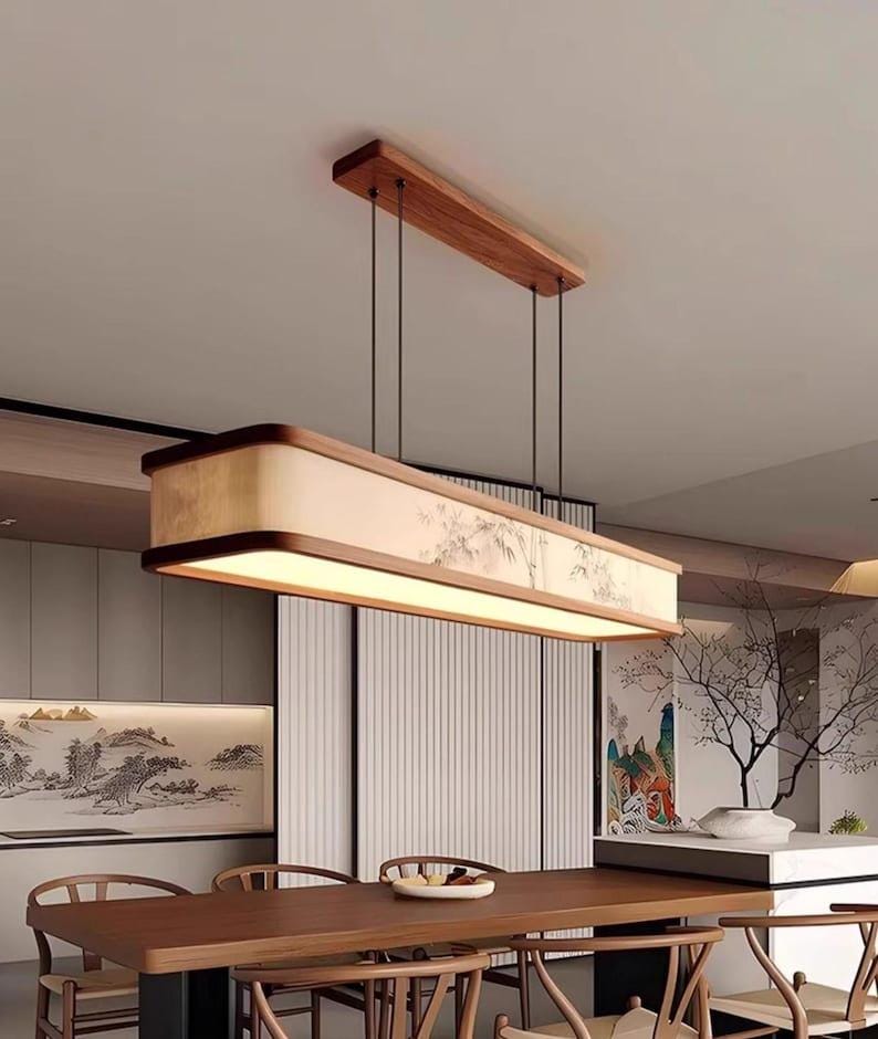 May include: A rectangular, wooden pendant light fixture with a warm, ambient glow. The fixture is suspended from the ceiling by thin black cords and features a light-colored shade with a subtle, Asian-inspired landscape design. The fixture is above a wooden dining table.