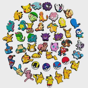 Op de afbeelding: Een verzameling kleurrijke cartoon karakter bedels, waaronder Pikachu, gerangschikt in een cirkelvormig patroon. De bedels hebben verschillende ontwerpen en kleuren, sommige lijken op ballen. De achtergrond is effen lichtgrijs.