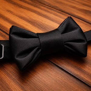 May include: A close-up of a black bow tie on a wooden surface. The bow tie is made of a smooth, satin-like material and features a classic butterfly shape. The adjustable strap and metal clasp are visible.