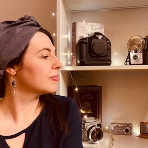 May include: A woman wearing a gray headband and dangling earrings. A black camera, flash, and other vintage cameras are on a shelf in the background. The woman is wearing a dark blue top.