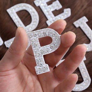 Puede incluir: Una letra "P" blanca con brillantes pedrería se sostiene en una mano. La letra forma parte de un conjunto de letras decorativas, con otras visibles en el fondo. Los brillantes transparentes están dispuestos sobre la superficie de la letra.