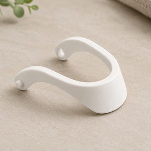 May include: A white, U-shaped plastic component featuring a smooth, matte finish. The object has two curved arms, one ending in a small circular hole and the other featuring a small protruding peg, designed for a mechanical connection.