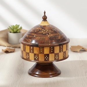 Op de afbeelding: Een decoratieve houten doos met een gewelfd deksel en een voetstuk. De doos heeft een dambordpatroon rond de basis en is versierd met ingelegde messing accenten. Het deksel is bekroond met een kleine bekroning.