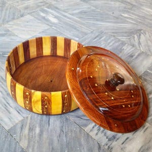 May include: A round wooden bowl with a lid. The bowl's rim features alternating vertical stripes of light and dark wood. The lid is oval-shaped with a dark knob and decorative inlaid patterns. The bowl is open, revealing a smooth, dark interior.