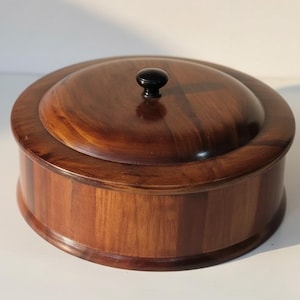 May include: A round wooden box with a lid. The box is made of dark brown wood with a polished finish. The lid has a small, black knob. The box is likely used for storage or display.