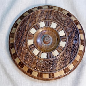 May include: A circular wooden plate with detailed floral carvings. The plate has a dark brown central area, a lighter wood border, and accents. The design includes a raised central section and a detailed floral pattern around the edge.