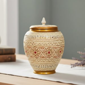 May include: A decorative beige and gold ceramic jar with a lid. The jar features intricate patterns in red and gold, with a gold base and rim. The lid is topped with a white finial. The jar is likely used for storage or decoration.