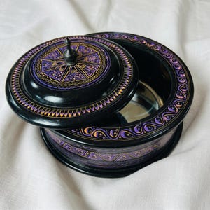 May include: A black, round wooden box with a lid, featuring intricate purple and gold painted designs. The lid is slightly ajar, revealing the interior. The box sits on a white surface.