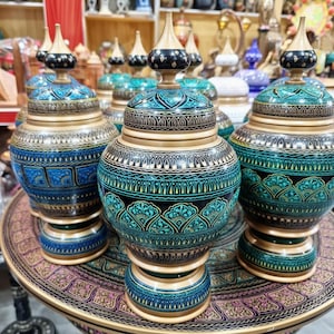 Op de afbeelding: Handgemaakte houten vazen met deksels. De vazen hebben ingewikkelde patronen in teal, goud en zwart. De deksels zijn afgewerkt met een zwarte en gouden top. De vazen worden tentoongesteld op een decoratieve tafel.