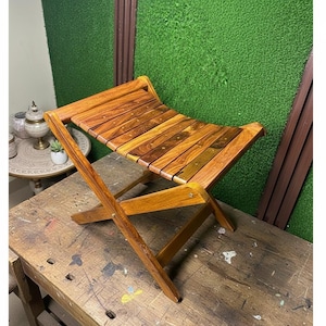 May include: A folding wooden stool with a slatted seat. The stool is made of warm-toned wood and has a simple, functional design. The legs are crossed for stability. The stool is suitable for indoor or outdoor use.