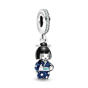 May include: A silver charm featuring a Kokeshi doll. The doll has a black bob and wears a blue kimono with white floral patterns. The charm has a ring with the word "Pandora" and small clear stones.