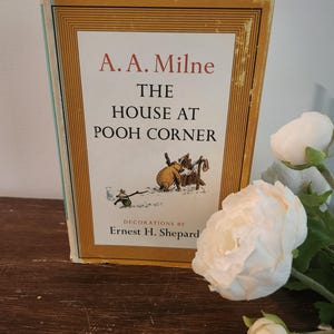 May include: A hardcover book titled The House at Pooh Corner by A. A. Milne with decorations by Ernest H. Shepard. The cover features a cream background with a gold-lined border and a small illustration of Winnie the Pooh and Piglet.