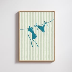 Puede incluir: Impresión enmarcada de un bikini turquesa colgado de un tendedero. El fondo tiene rayas verticales en verde claro y blanco. La obra de arte tiene un estilo minimalista, ideal para la decoración de playa o verano.