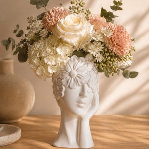 Puede incluir: Un jarrón de cerámica blanca con forma de cabeza de mujer, adornado con detalles florales, contiene un ramo de flores. El arreglo incluye una gran rosa color crema, claveles melocotón y acentos blancos y verdes.