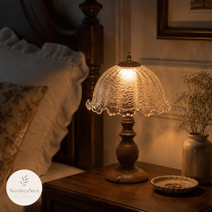 May include: A table lamp with a dark brown wooden base and a clear, crackled glass shade featuring a scalloped edge. The lamp emits a warm, glowing light from within the textured glass dome, which is supported by a turned wooden pedestal stand.