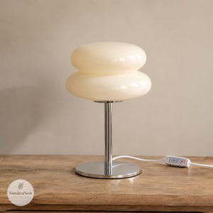 May include: A table lamp featuring a double-tiered, cream-colored glass shade with a smooth, rounded profile. The lamp is supported by a polished chrome metal stem and a circular base, accompanied by a white power cord with a control switch.
